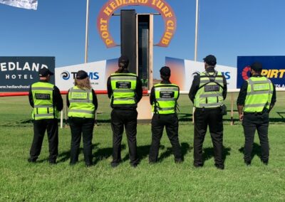 6 Security Guards in Hi-Viz standing infront of a sign saying "Port Hedlan Turf Club" facing away from the camera